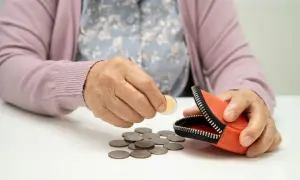 Dłoń starszej osoby wkłada monetę do portmonetki. Wokół leżą rozsypane monety, symbolizujące oszczędności lub potrzebę wsparcia, np. z PFRON dofinansowanie.