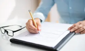 Dłoń pisze na dokumencie, być może wniosek o sudop pomoc de minimis. Obok leżą okulary.