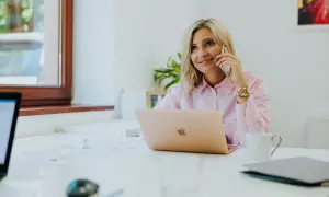 Kobieta w różowej koszuli rozmawia przez telefon przy biurku z laptopem.