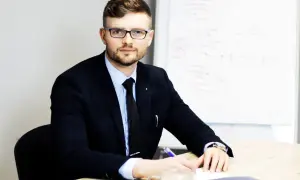 Mężczyzna w garniturze i okularach siedzi przy biurku, pisząc coś na kartce. Obok filiżanka kawy.