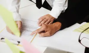 Dłonie dwóch osób nad dokumentem, jedna pisze, druga wskazuje palcem.