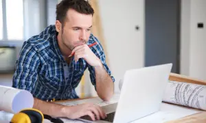 Mężczyzna w kratę pracuje na laptopie, trzymając ołówek, obok plany i słuchawki.