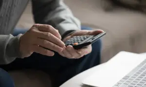 Dłonie mężczyzny liczą na kalkulatorze, planując swój kapitał własny. Obok leży laptop.