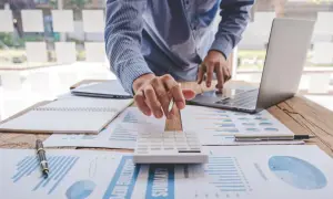 Osoba przy biurku z laptopem, kalkulatorem i dokumentami finansowymi.