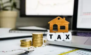 Drewniany domek na kostkach z napisem TAX, stosy monet i wykresy. Symbolizuje podatek od wzbogacenia i inwestycje.