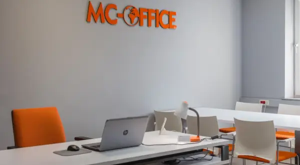 Pomarańczowe logo "MC-OFFICE" na szarej ścianie, biurko z laptopem i krzesło.