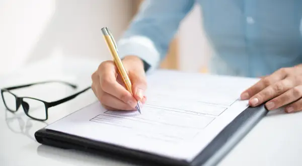 Dłoń pisze na dokumencie, być może wniosek o sudop pomoc de minimis. Obok leżą okulary.