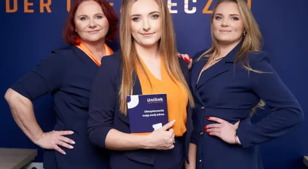 Trzy kobiety w granatowych marynarkach i pomarańczowych bluzkach pozują na tle niebieskiej ściany z logo.
