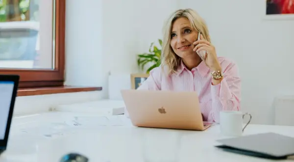Kobieta w różowej koszuli rozmawia przez telefon przy biurku z laptopem.