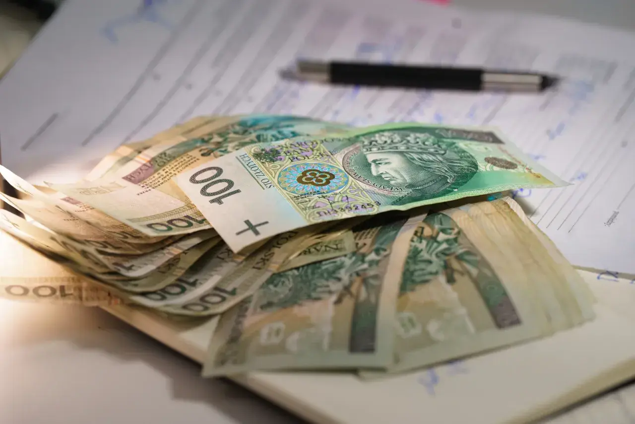 Stos stu-złotowych banknotów leży na dokumentach. W tle widać długopis. To może być pomoc dla osób z mops znaczny stopień niepełnosprawności.