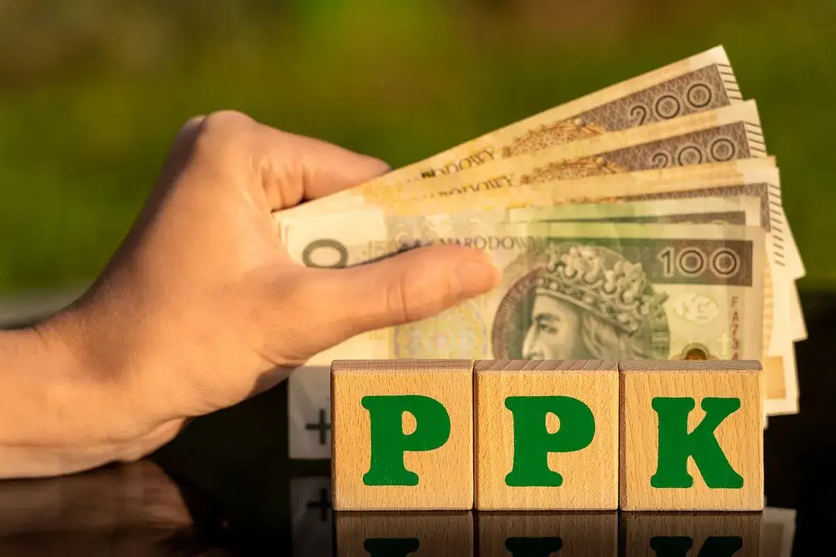 Dłoń trzyma banknoty, obok kostki z literami PPK. To symbol pracowniczego planu kapitałowego, który buduje Twoją przyszłość finansową.
