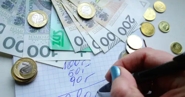 Ręka z niebieskim lakierem pisze na kratkowanym papierze, obok leżą polskie banknoty i monety. Planowanie wydatków to pierwszy krok do rewaloryzacji budżetu.